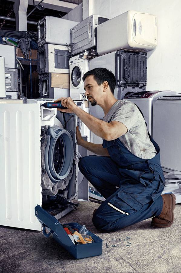 Un technicien répare un lave-linge dans un atelier. Il porte une tenue de travail bleue et utilise un tournevis. À l'arrière-plan, on peut voir d'autres appareils électroménagers.