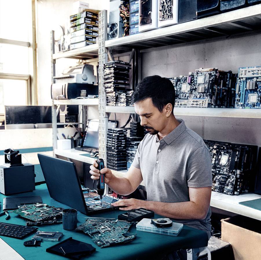 Un homme répare des pièces d'ordinateur sur une table, entouré d'étagères remplies de composants électroniques dans un atelier.