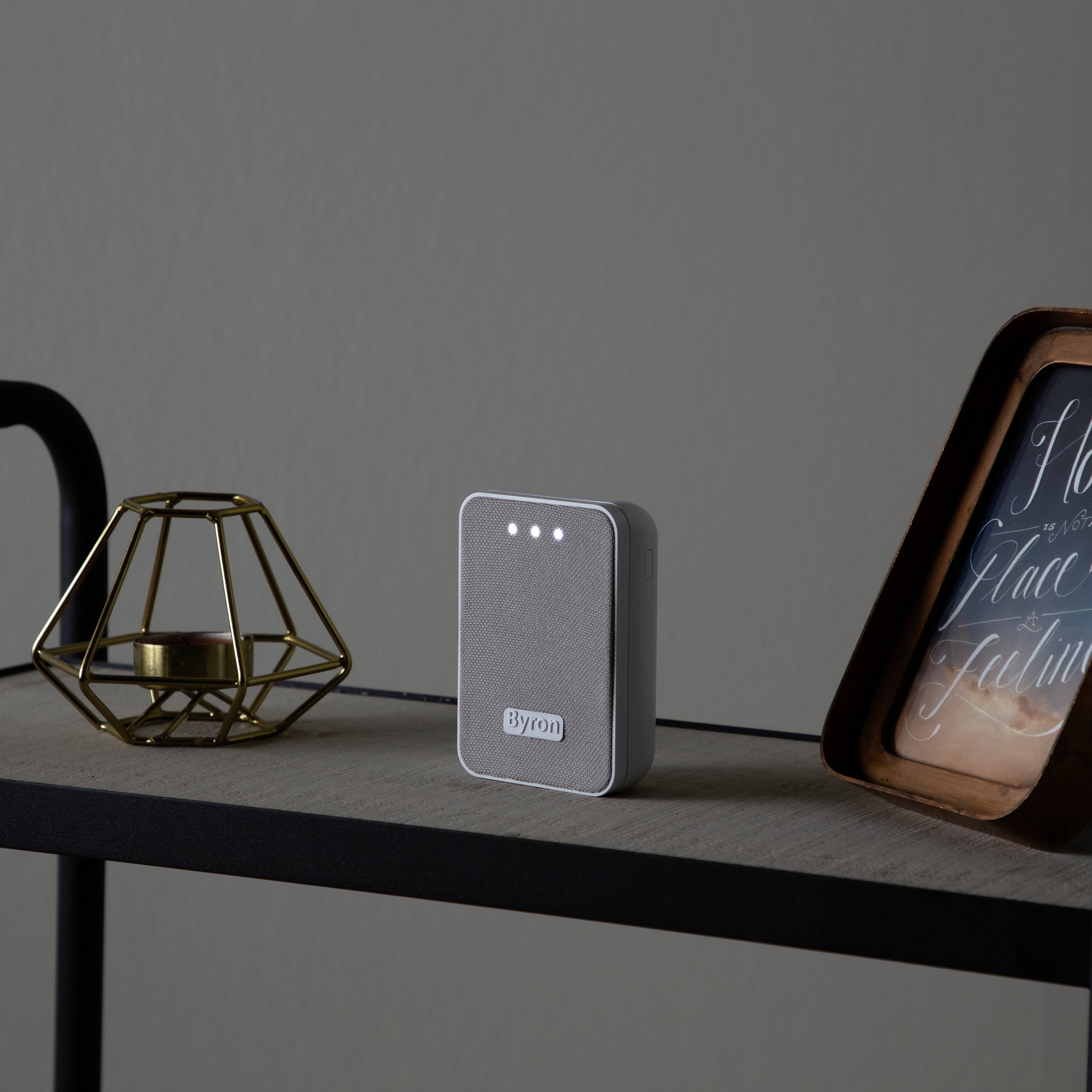 Un appareil gris et rectangulaire avec trois voyants lumineux en haut est posé sur une table, à côté d'un bougeoir doré et d'une image encadrée.