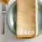 Serviette en tissu jaune portant le nom 'Tara' sur une assiette verte, à côté d'une fourchette.