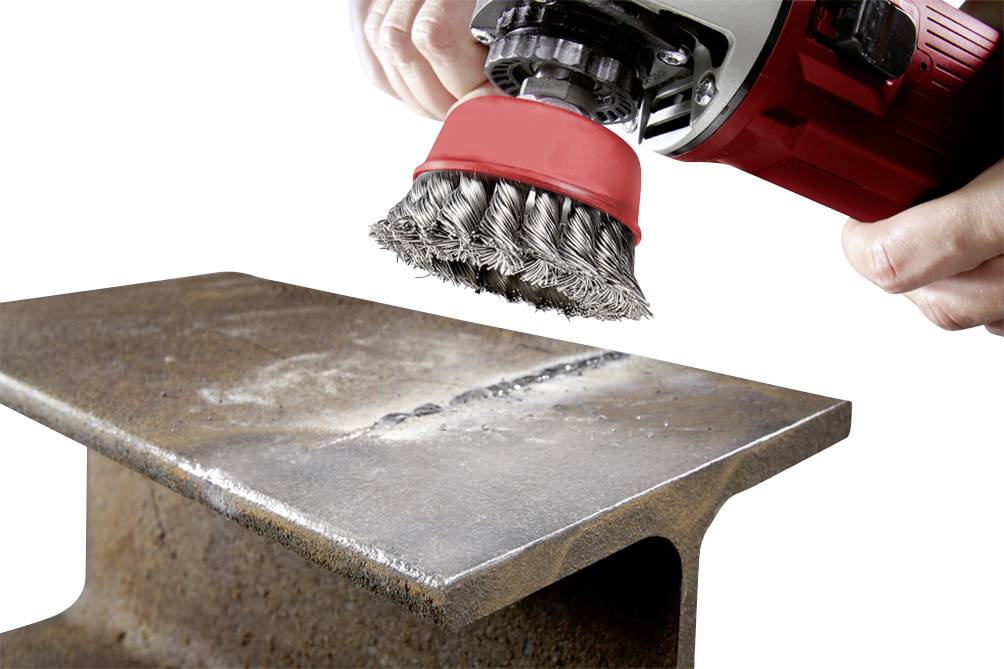 Une main tient une brosse métallique rouge et noire fixée à une meuleuse d'angle pour enlever la rouille d'une poutre métallique.