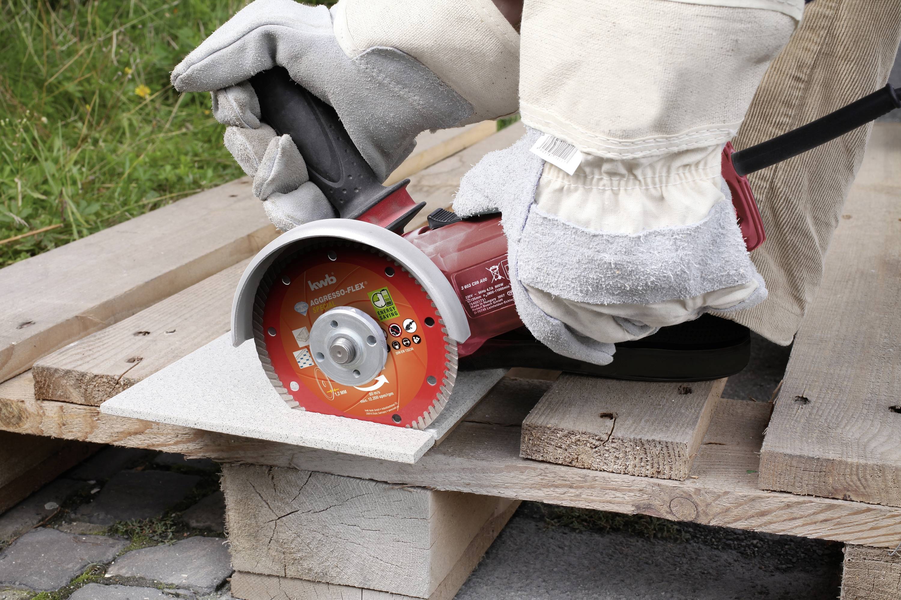 Une personne scie une dalle de céramique grise sur des palettes en bois à l'aide d'une scie circulaire rouge. La personne porte des gants de protection et un pantalon de travail.