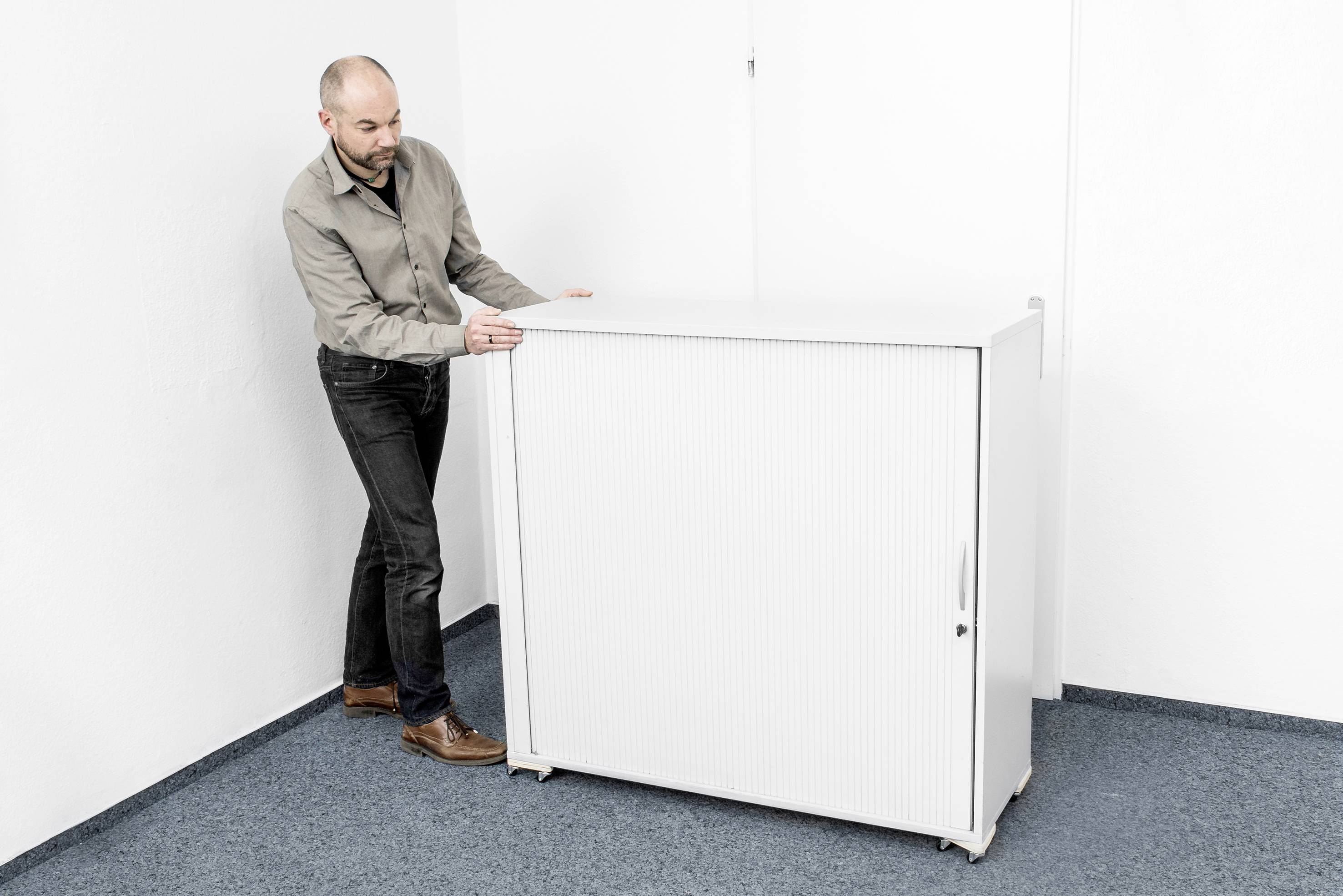 Un homme en chemise grise et jeans pousse une armoire de bureau blanche sur un sol recouvert d'un tapis bleu contre un mur blanc.