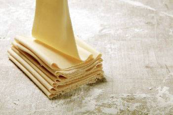 Une pile de feuilles de lasagne fraîches et non cuites est posée sur une table farinée. Une feuille est en train d'être retirée de la pile.