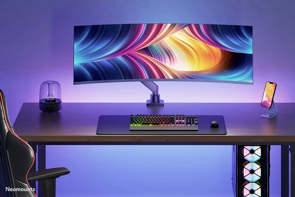 Moniteur ultra-large avec un écran coloré sur un bureau, clavier rétroéclairé et support de téléphone à côté, lumière ambiante.