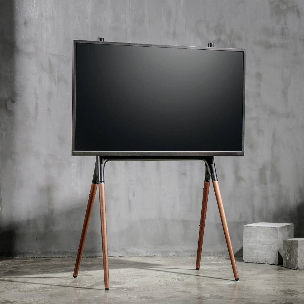 Un téléviseur moderne est posé sur un support en bois à trois pieds dans une pièce minimaliste aux murs en béton.