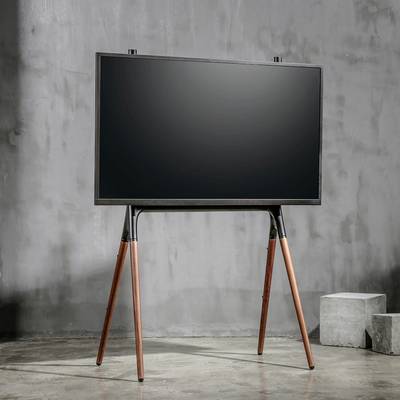 Un téléviseur moderne est posé sur un support en bois à trois pieds dans une pièce minimaliste aux murs en béton.
