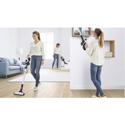 Une femme utilise un aspirateur sans fil en trois parties : elle aspire le tapis, nettoie une étagère, monte les escaliers. Environnement de salon lumineux et moderne.