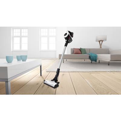 Aspirateur sans fil posé sur un parquet dans un salon moderne avec canapé, table et lampe. Intérieur lumineux et minimaliste.