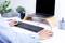 Une personne travaille à un bureau avec un ordinateur portable, un clavier et une souris. L'ordinateur portable est posé sur un support en bois. Une plante et une tasse sont placées à côté.