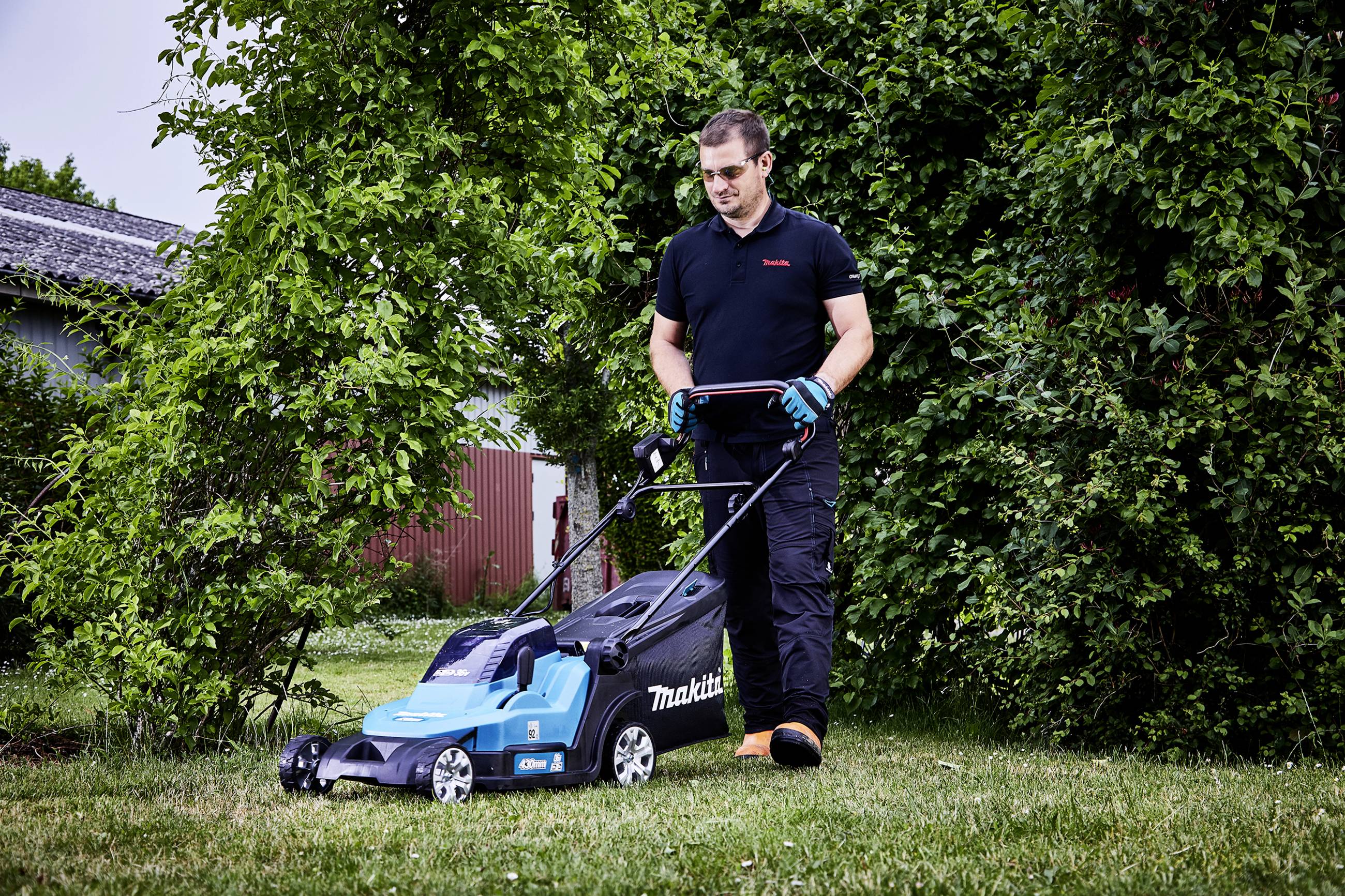 Un homme tond la pelouse avec une tondeuse Makita bleue dans un jardin avec des buissons verts en arrière-plan.