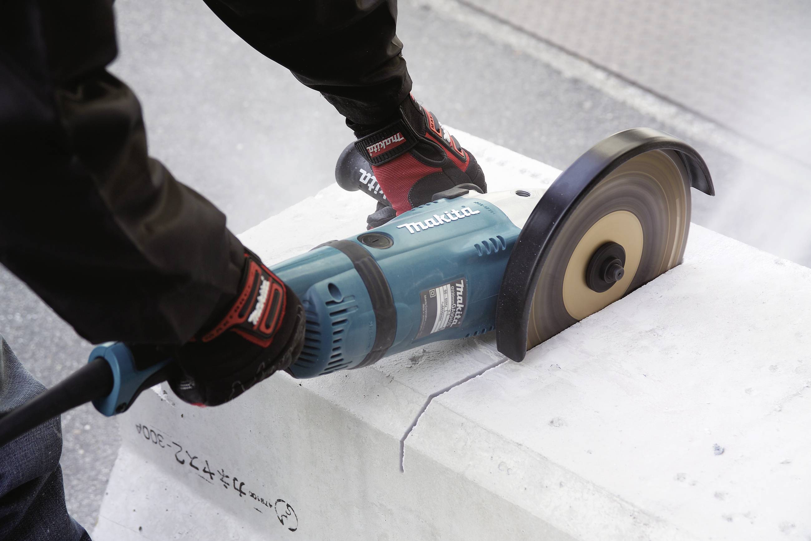 Une personne coupe un bloc de béton avec une scie circulaire portative, portant des gants et des vêtements de protection ; de la poussière est soulevée.
