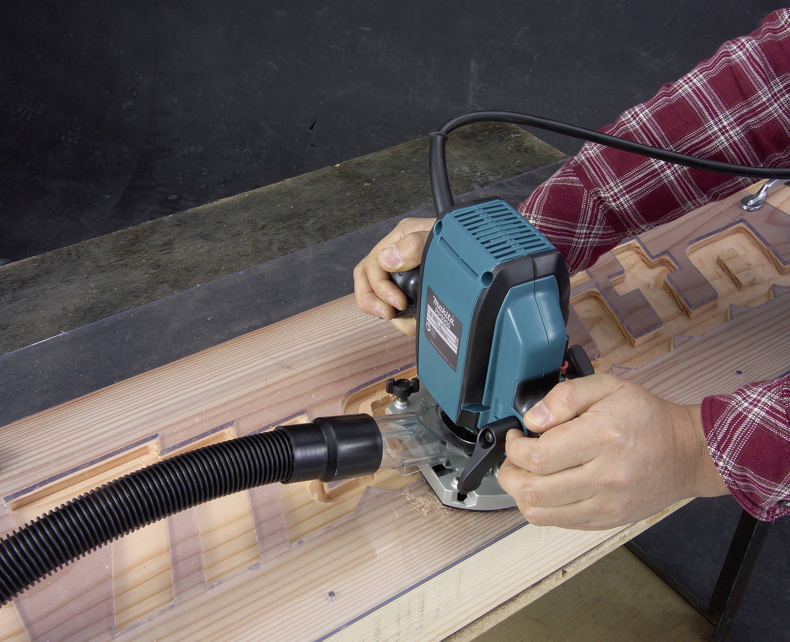 Une personne utilise une défonceuse pour travailler le bois. Les copeaux sont aspirés par un tuyau. Des lettres sont visibles sur le bois.