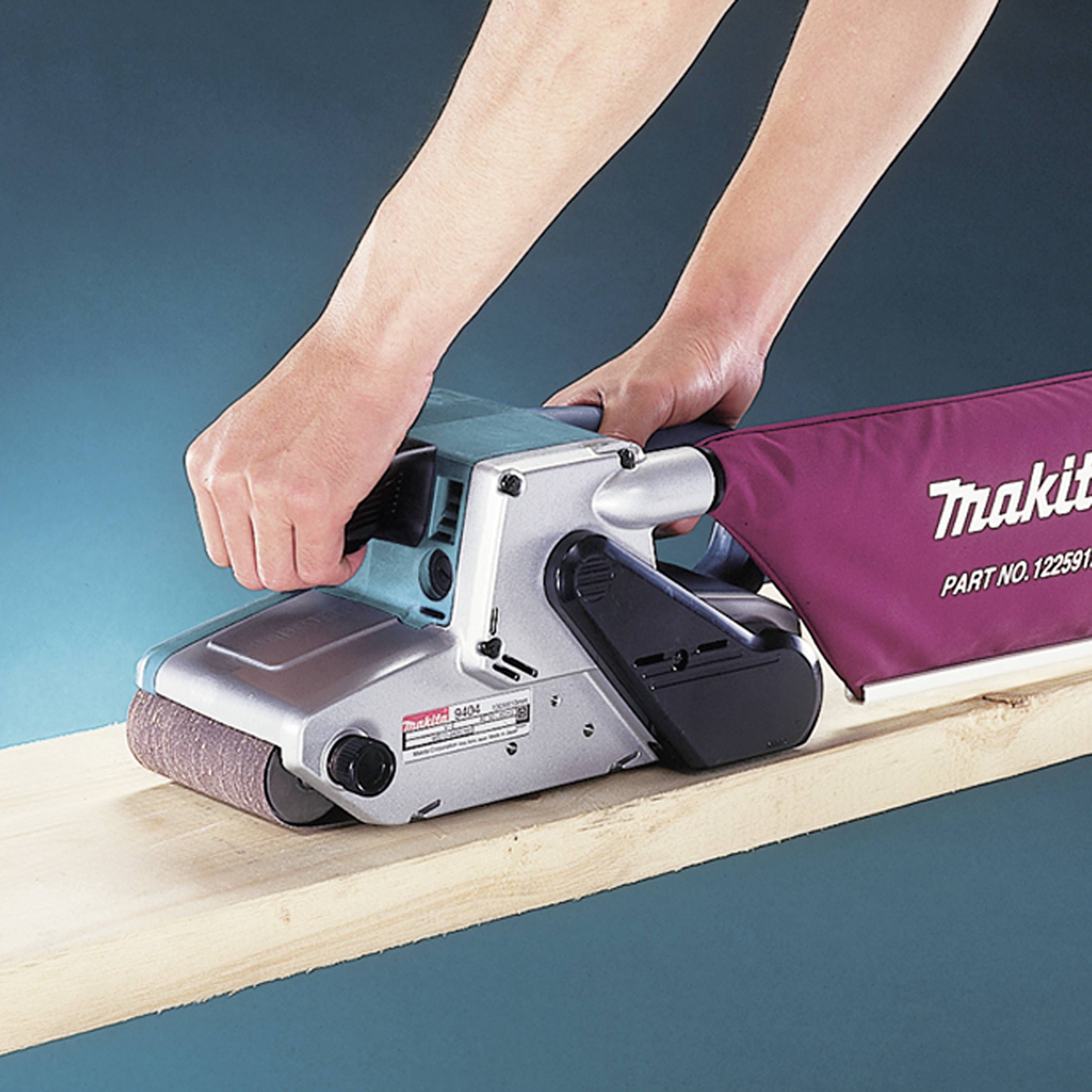 Une personne utilise une ponceuse pour faire du poncage sur une planche en bois. La ponceuse a un sac de récupération de poussière rouge.