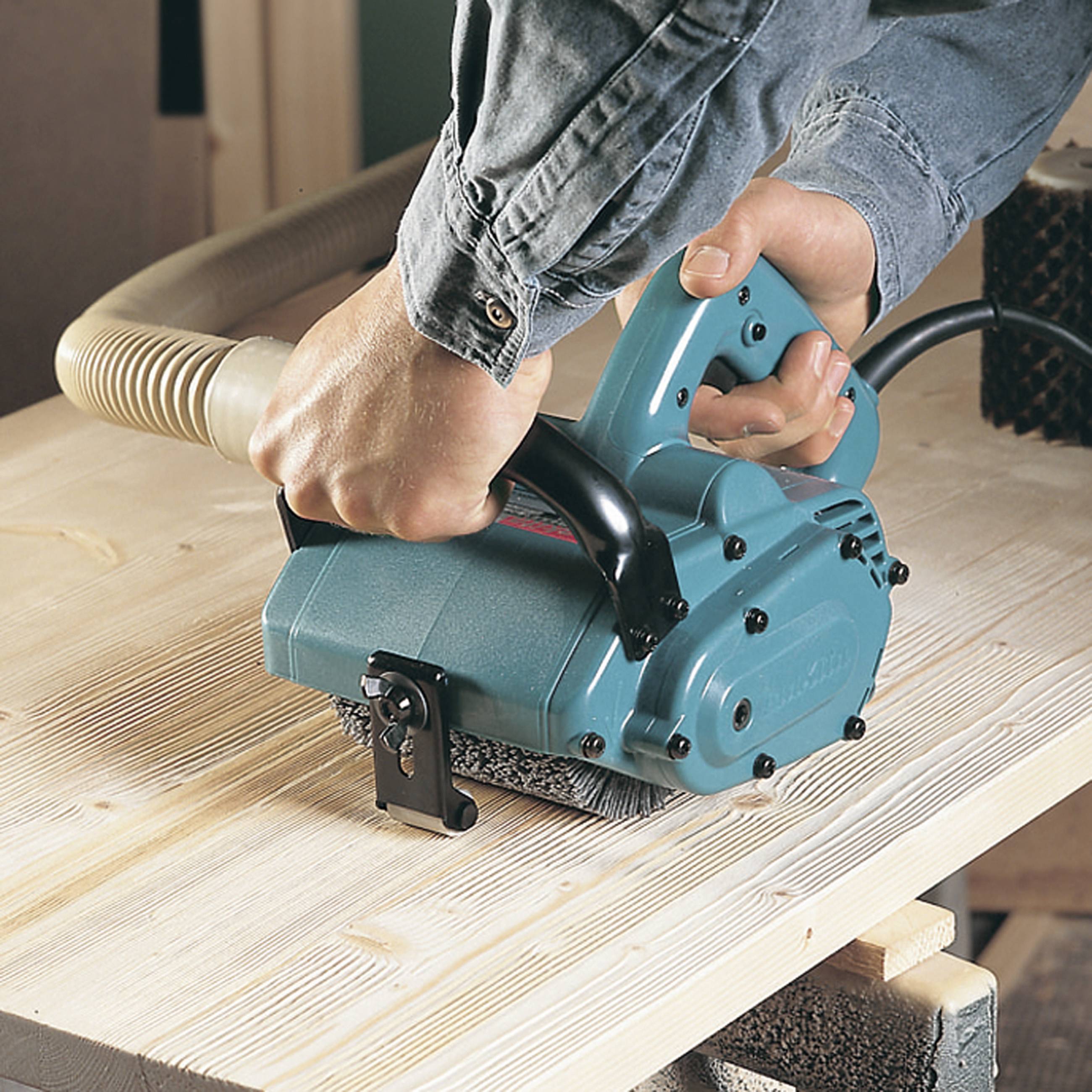 Une personne utilise un rabot électrique pour lisser une surface en bois. Un tuyau d'aspiration enlève les copeaux pendant le rabotage.