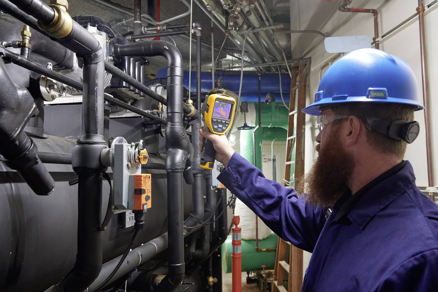 Un technicien en tenue de protection et casque utilise une caméra thermique pour inspecter des tuyaux industriels dans une salle de machines.