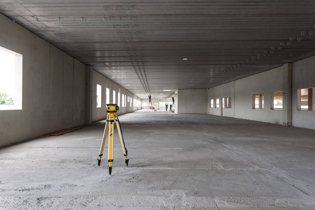 Un instrument de topographie est placé dans une grande pièce vide et inachevée, au sol en béton et aux plafonds hauts. Des ouvriers sont visibles en arrière-plan.