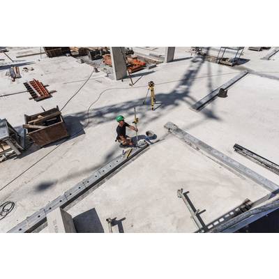 Un ouvrier sur un grand chantier, portant un casque vert, travaille avec un outil. Autour de lui, des poutres en acier et des matériaux de construction sont disposés.
