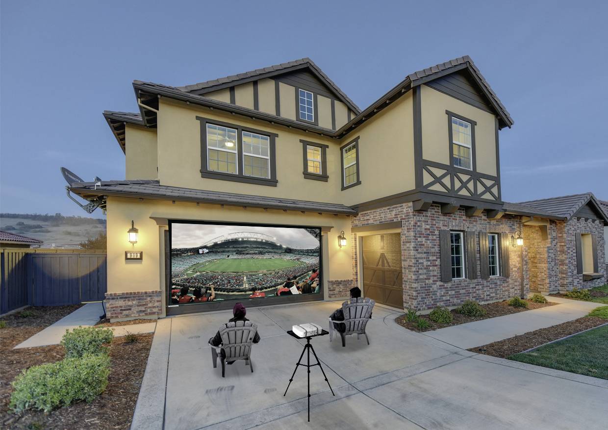 Deux personnes sont assises sur l'allée d'une grande maison et regardent un match de football sur un écran projeté dans le garage.