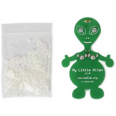 Une carte électronique verte d'alien portant l'inscription 'My Little Alien v1.0' à côté d'un sachet contenant de petits composants blancs.