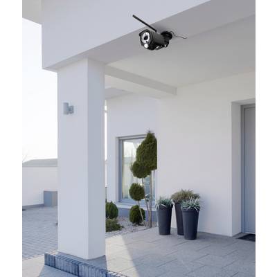 Caméra de surveillance montée sur un mur extérieur, montrant l'entrée avec deux plantes en pots et une fenêtre moderne.