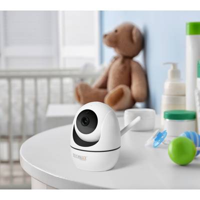 Une caméra pour bébé est posée sur une table dans la chambre d'enfant, à côté de jouets et de produits de soin. Un ours en peluche et un lit d'enfant sont visibles en arrière-plan.