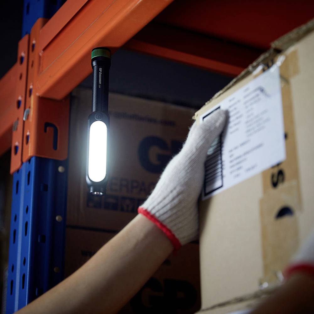 Une main gantée de blanc tient un carton dans un entrepôt. Une lampe torche est suspendue à une étagère et éclaire la scène.