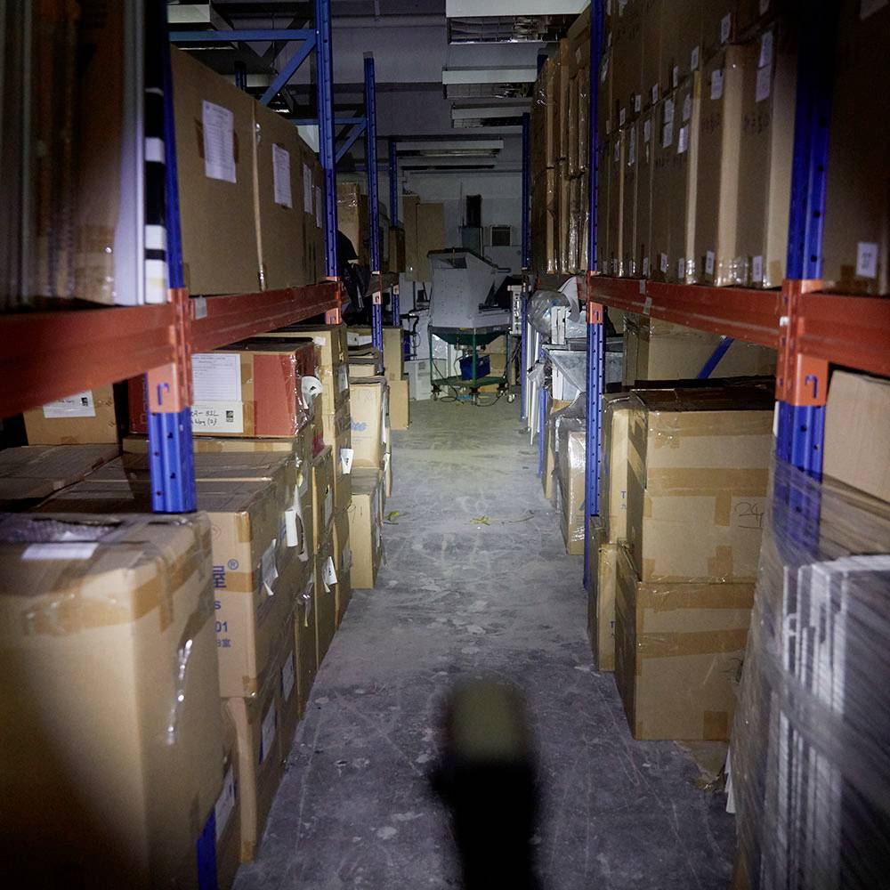 Un entrepôt avec des cartons empilés sur des étagères métalliques. Une machine se trouve à l'arrière-plan, et le sol est partiellement poussiéreux.