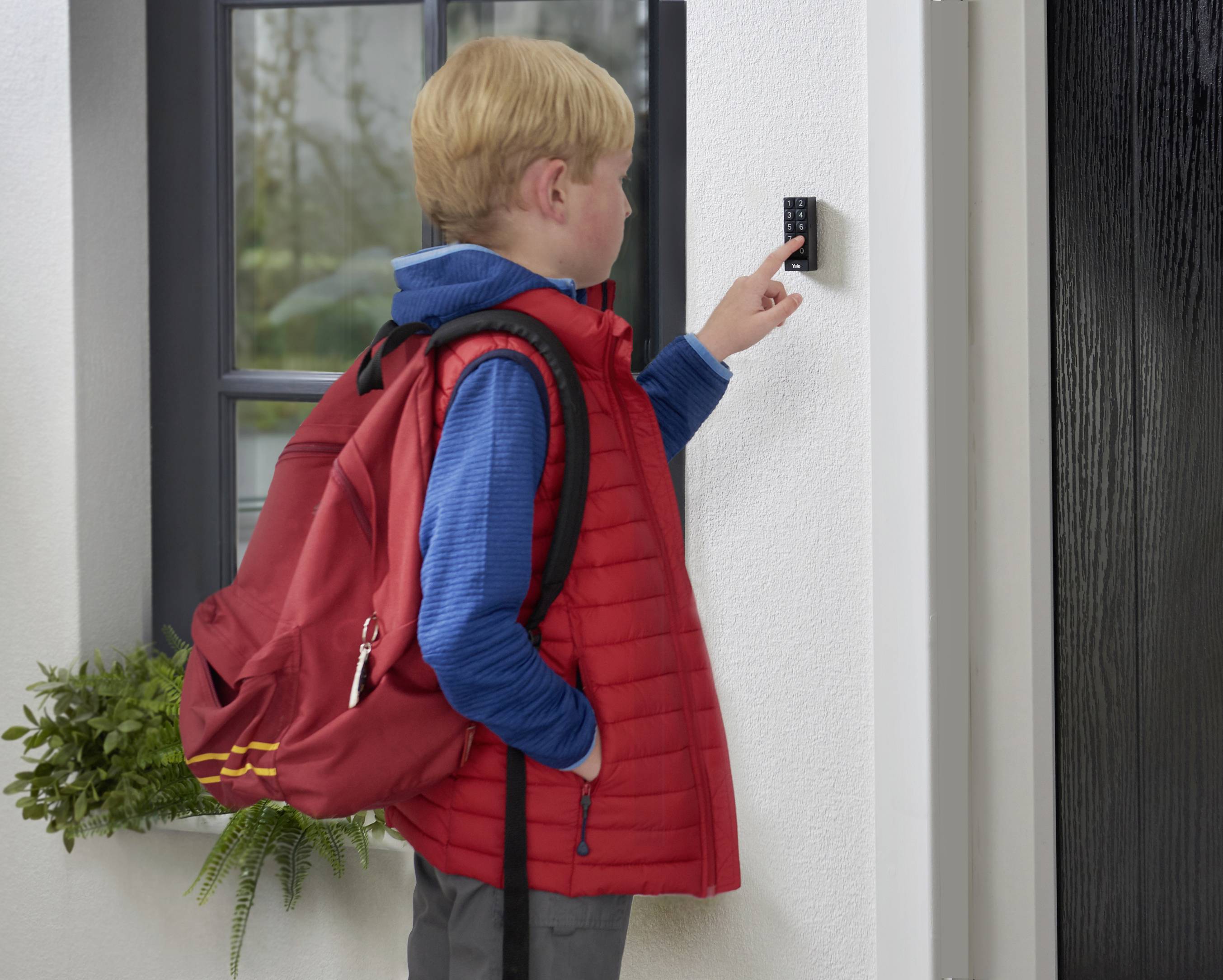 Un garçon avec un sac à dos rouge, un gilet rouge et une veste bleue sonne à une sonnette près d'une porte sur un mur blanc.