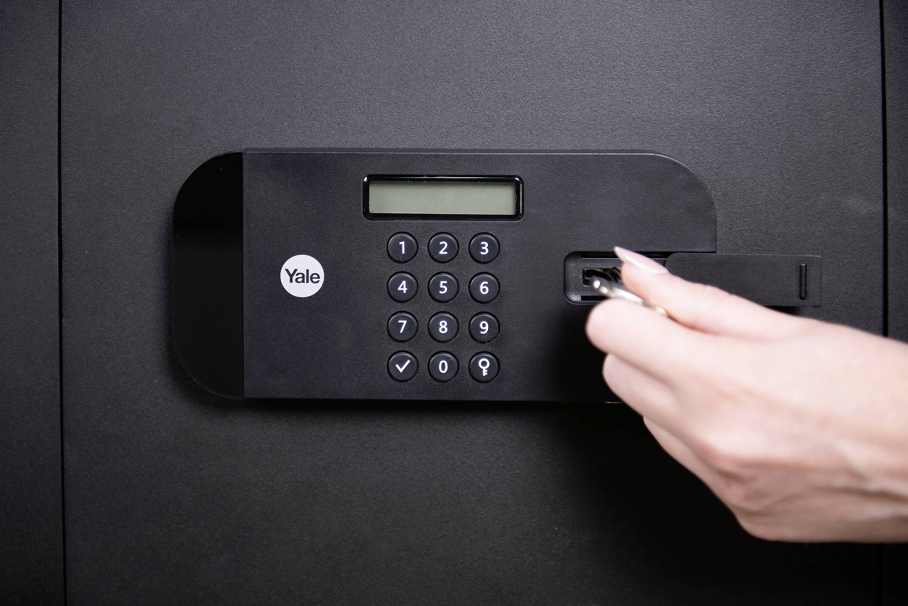 Une main tourne une clé dans la serrure d'un coffre-fort de sécurité équipé d'un clavier numérique et d'un écran LCD.