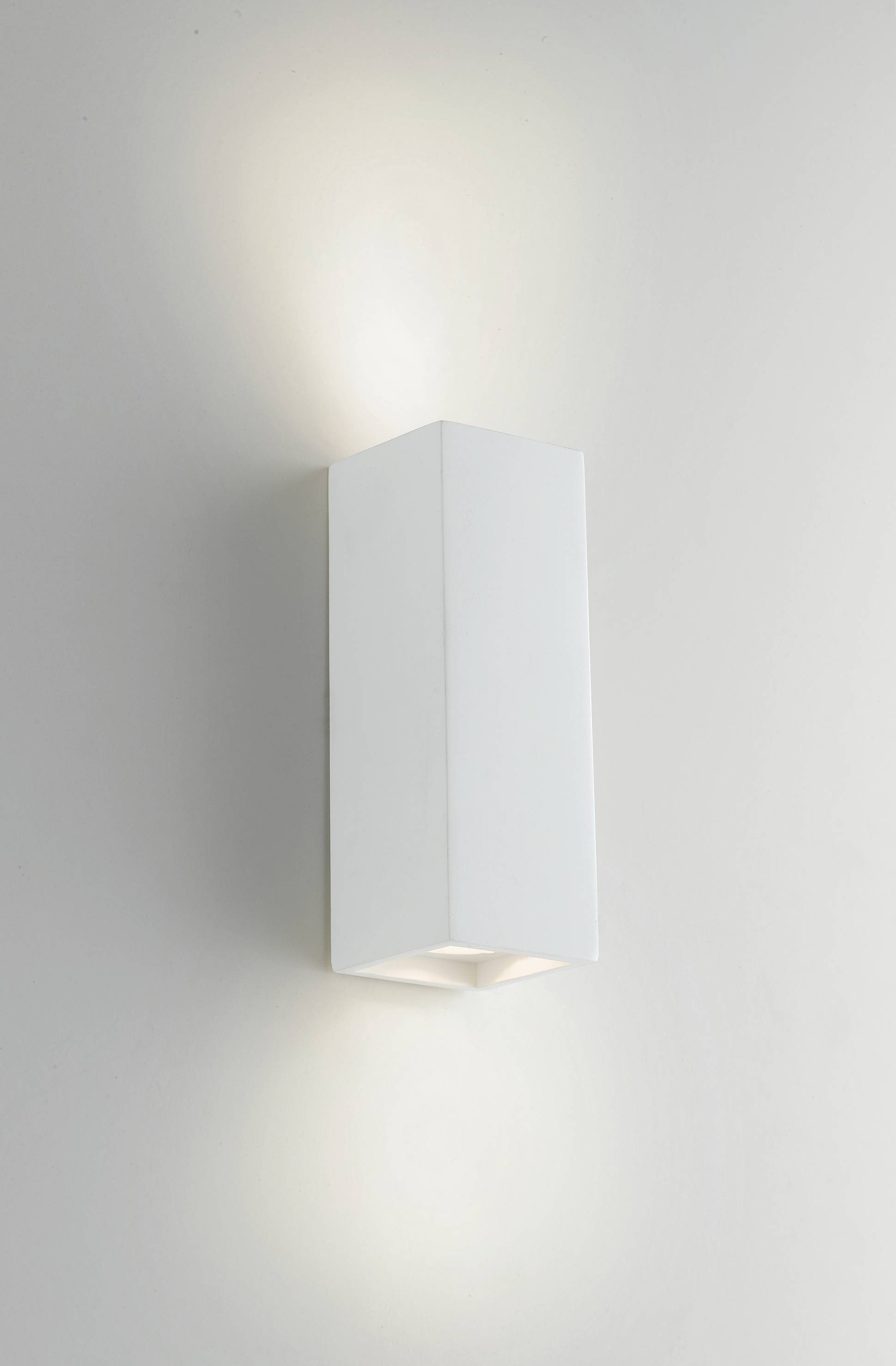 Applique murale blanche au boîtier rectangulaire. L'éclairage diffuse vers le haut et vers le bas, créant une ambiance douce sur le mur.