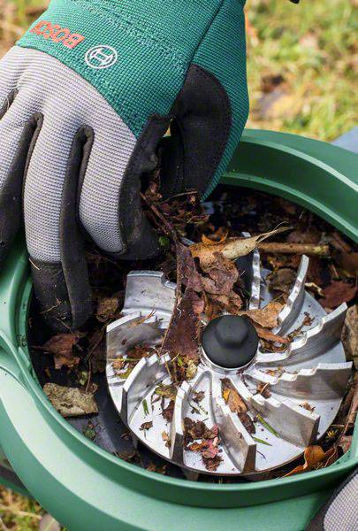 Une main gantée retire des débris de feuilles broyées d'un broyeur de jardin. Le rotor métallique est visible.
