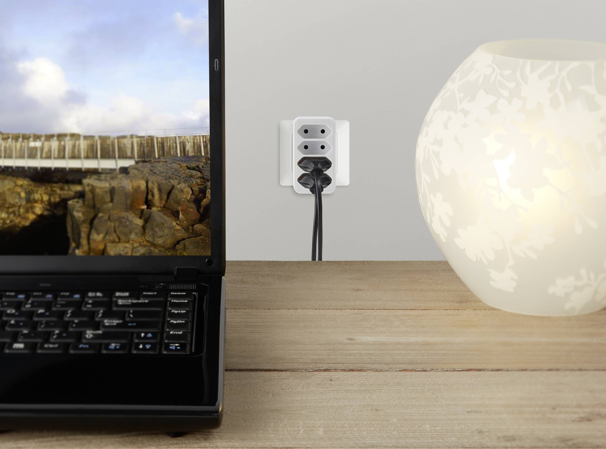 'Ordinateur portable, lampe et prise électrique sur une table en bois. L'ordinateur portable affiche un paysage côtier. La lampe est allumée, deux fiches sont branchées dans la prise.'