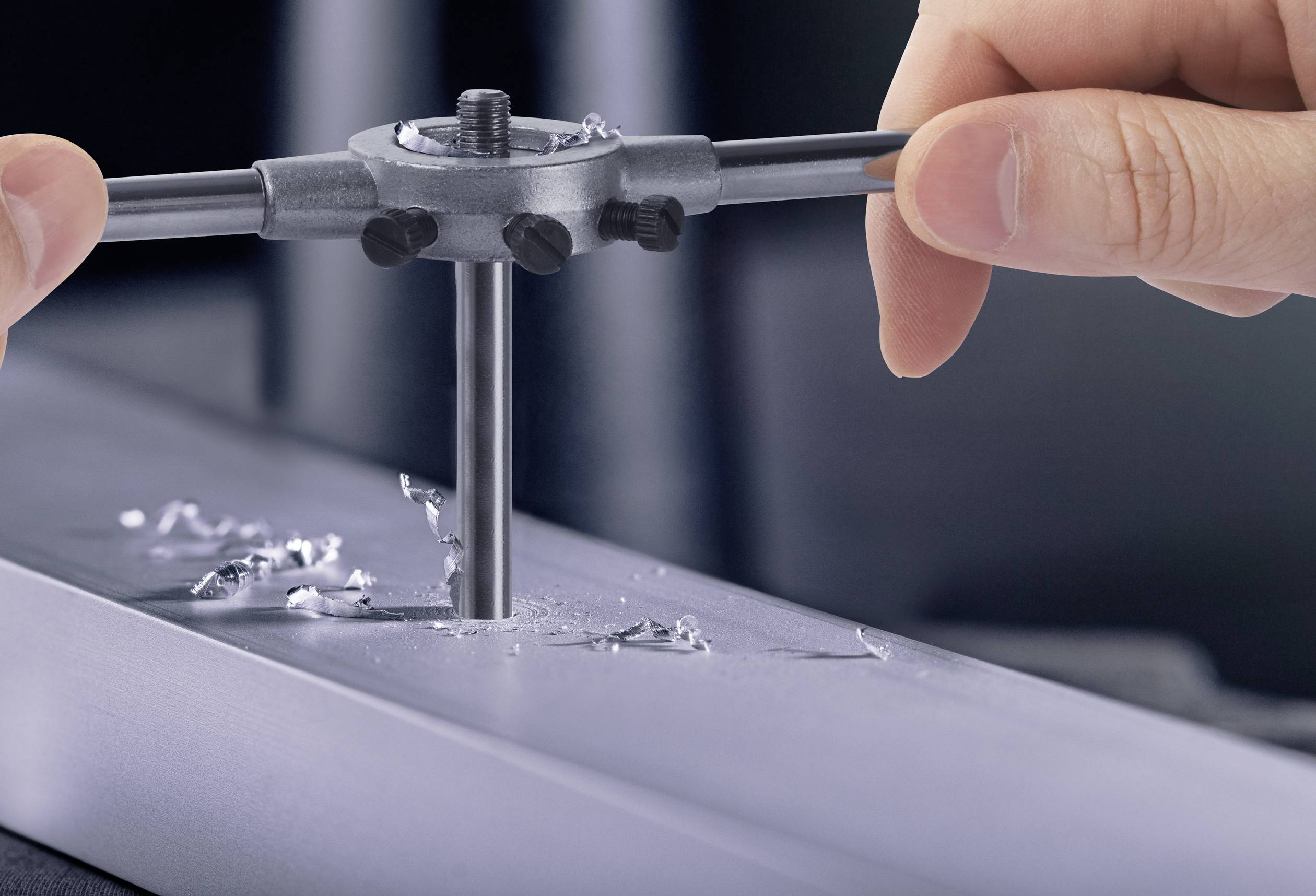 Deux mains utilisent un outil de filetage pour créer des filets précis dans un morceau de métal. Des copeaux de métal sont visibles.