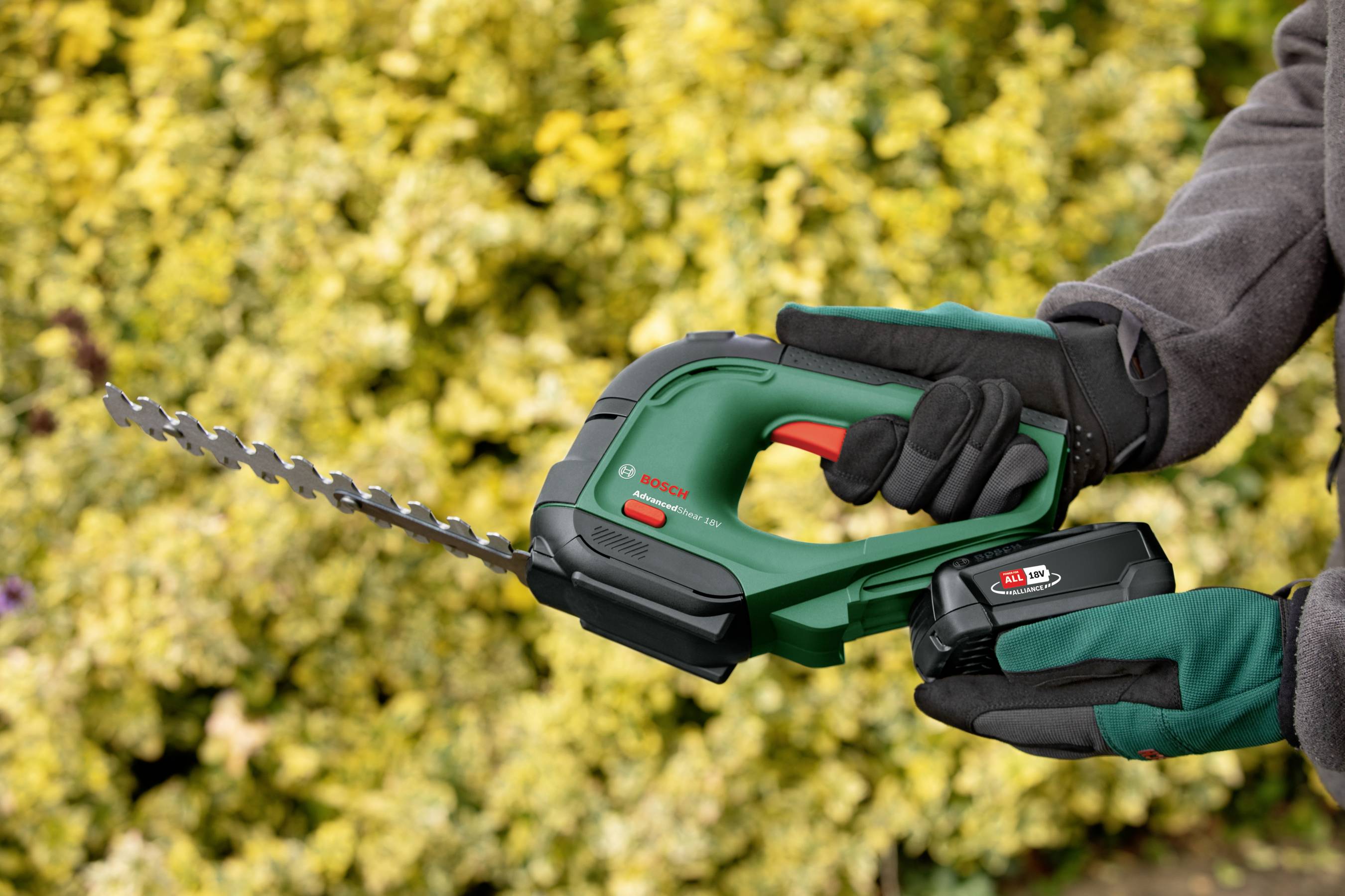 Une personne tient dans sa main une scie de jardin verte alimentée par batterie devant un fond de feuillage jaune.