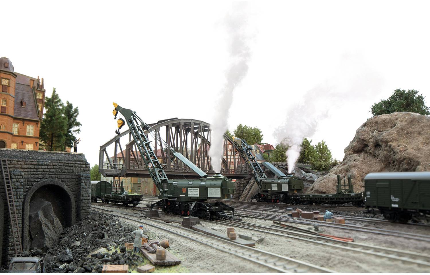 Scène de train miniature avec deux locomotives à vapeur, des grues et un tunnel, sur un fond de pont. Une figurine d'ouvrier au premier plan.