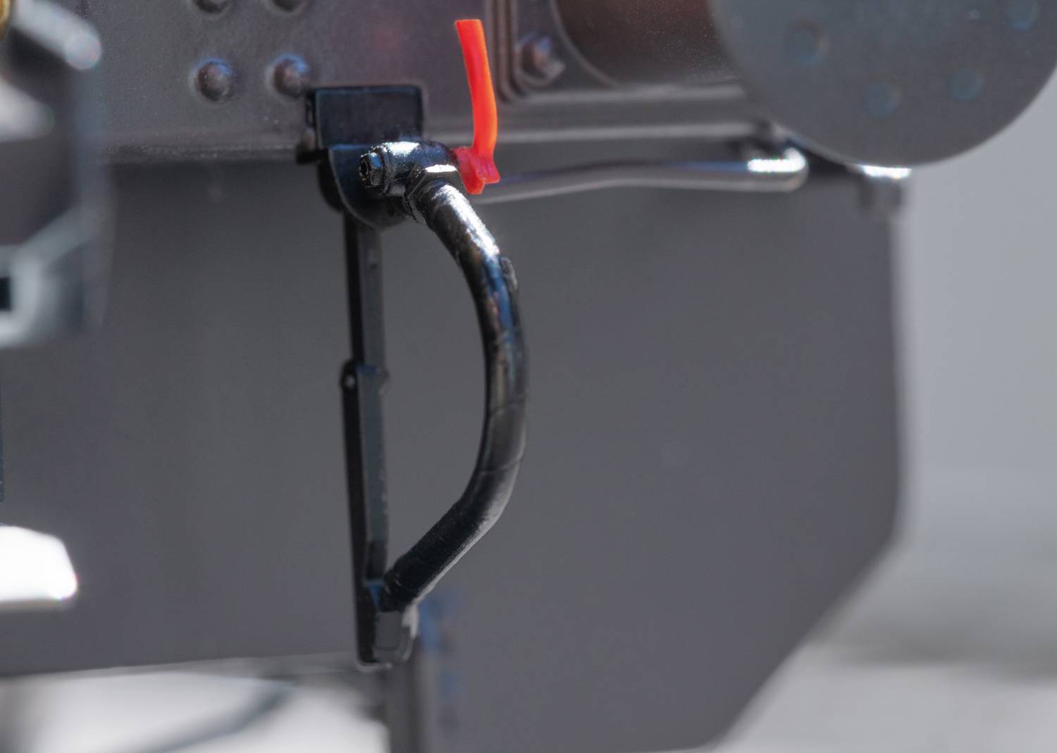 Une poignée noire en métal sur un dispositif mécanique, fixée à une surface grise. Un composant rouge en haut à droite.