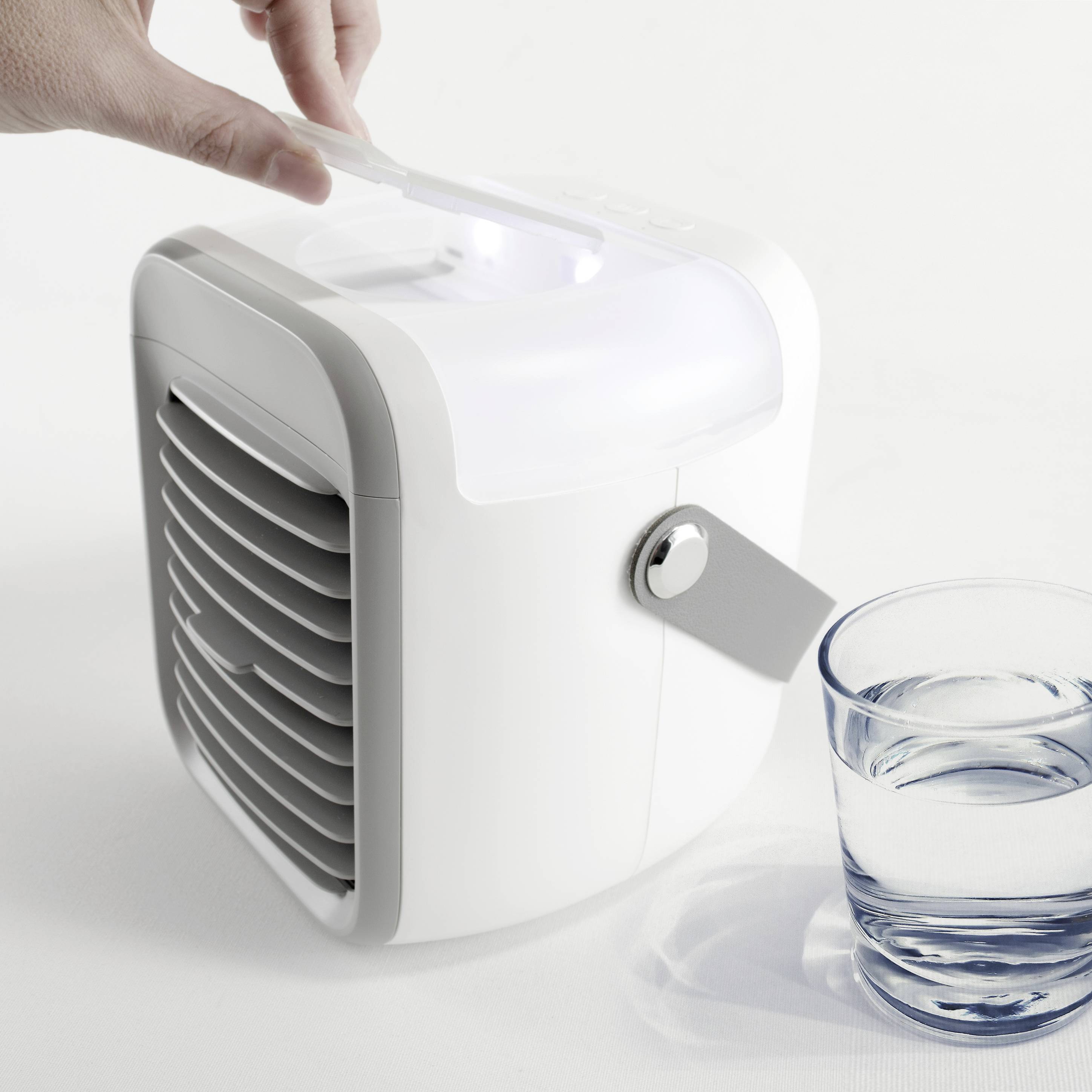 Un petit refroidisseur d'air à côté d'un verre d'eau. Une main effleure doucement le dessus du refroidisseur pour le faire fonctionner.