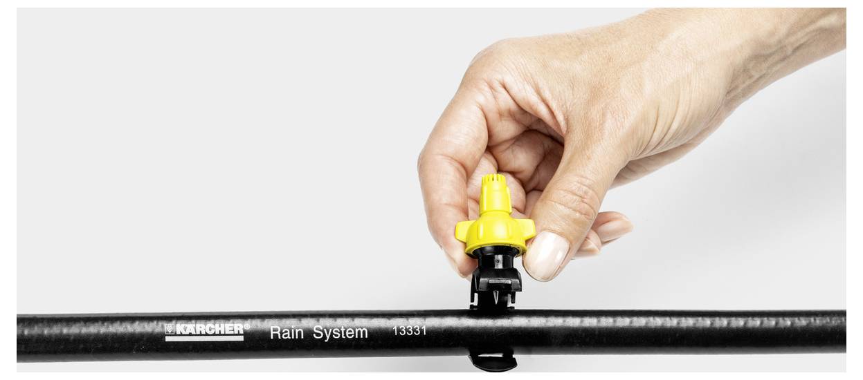 Une main ajustant un embout de goutte jaune sur un tuyau d'irrigation noir étiqueté « Système de pluie Karcher ».