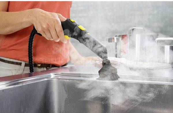 Une personne nettoie un évier de cuisine avec un nettoyeur vapeur. De la vapeur s'élève pendant que la brosse glisse sur la surface.