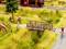 Un paysage miniature représente des voyageurs sur un chemin près d'un petit pont, avec un cheval dans un enclos et un cycliste.