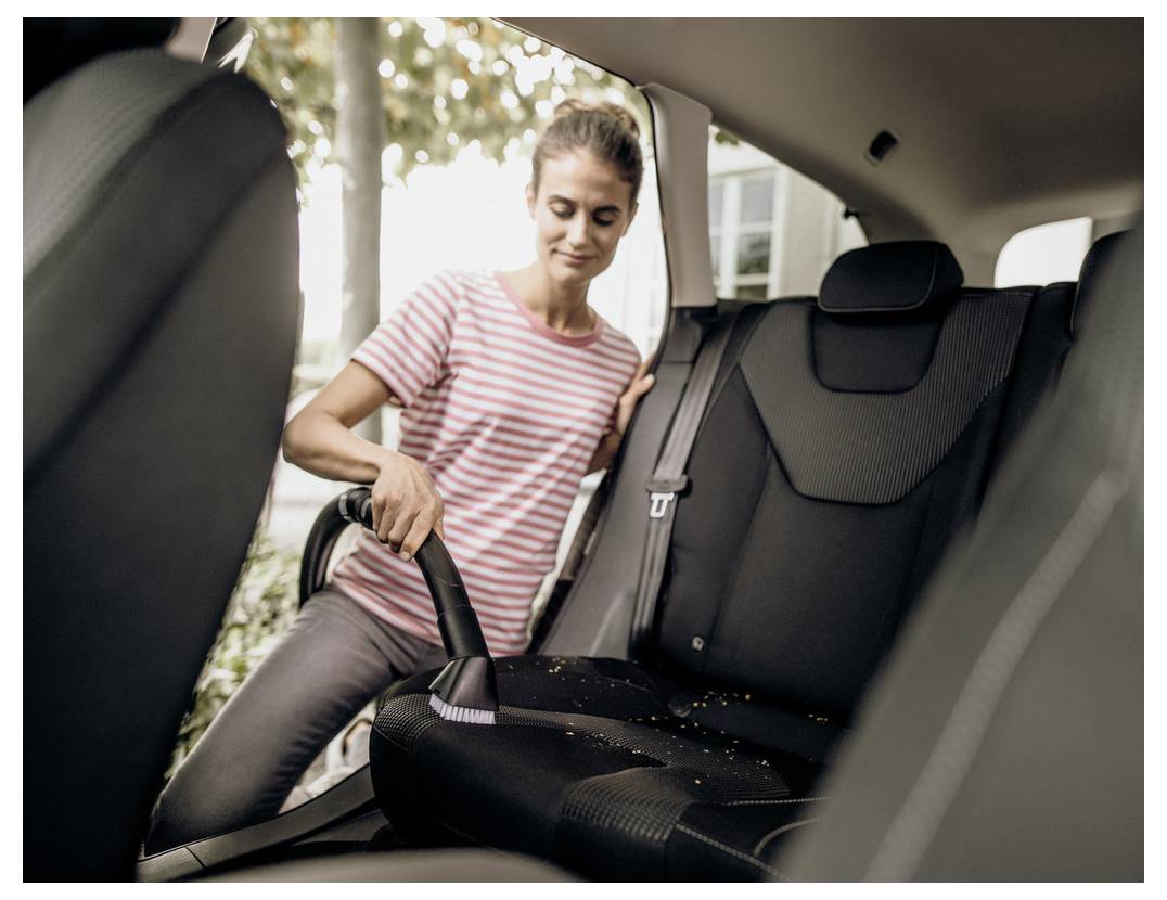 Une personne portant une chemise rayée est en train d'aspirer la banquette arrière d'une voiture, éliminant des débris. La lumière du soleil filtre à travers les vitres, suggérant une tâche de nettoyage.