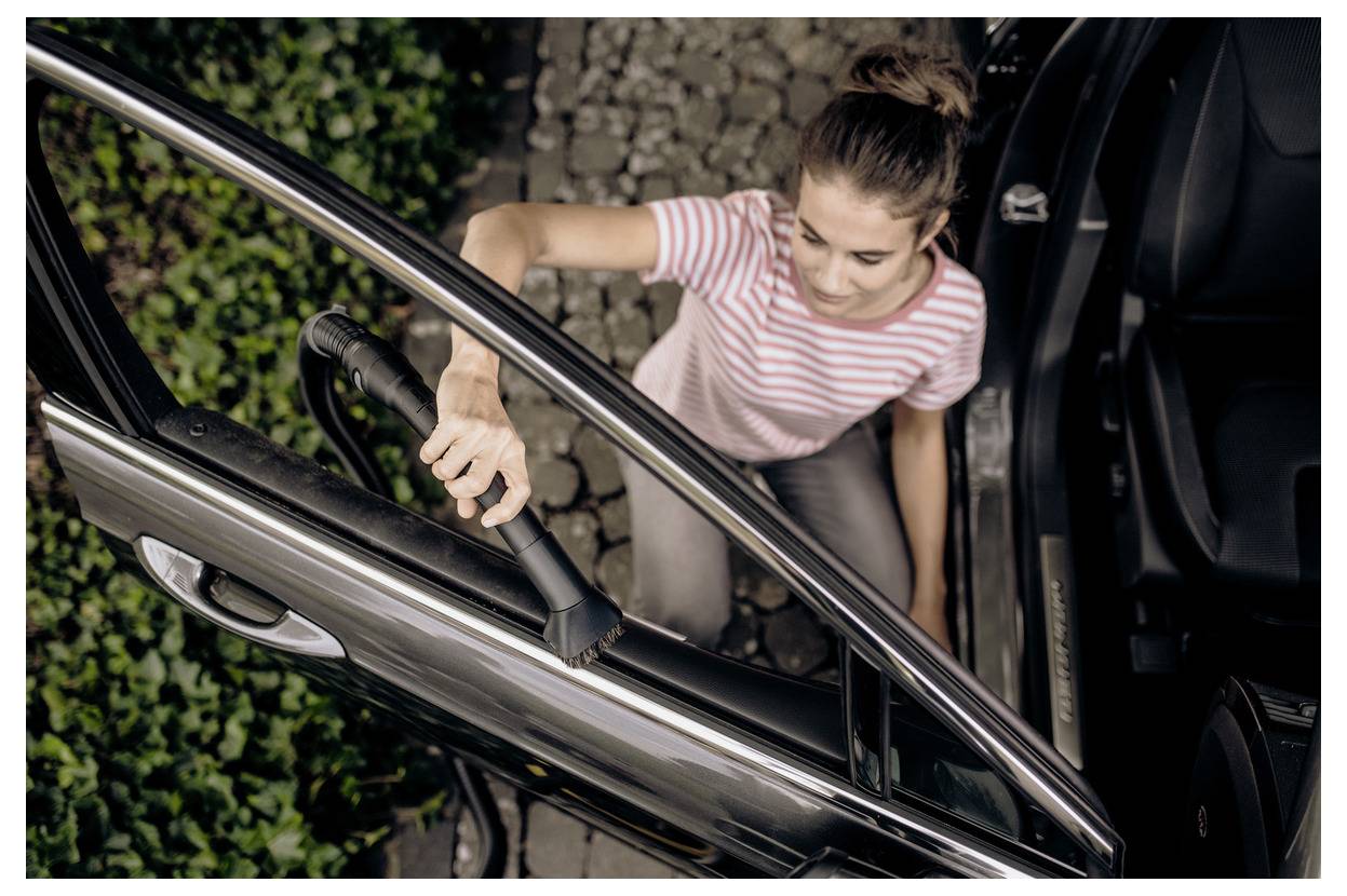 Femme portant un t-shirt rayé qui nettoie l'intérieur d'une voiture à l'aide d'un aspirateur sur une allée en pierre, vue de dessus.