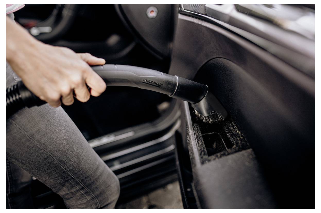 Une personne aspirant l'intérieur d'une voiture, concentrée sur la fissure entre le siège et le tableau de bord, à l'aide d'un aspirateur portable.