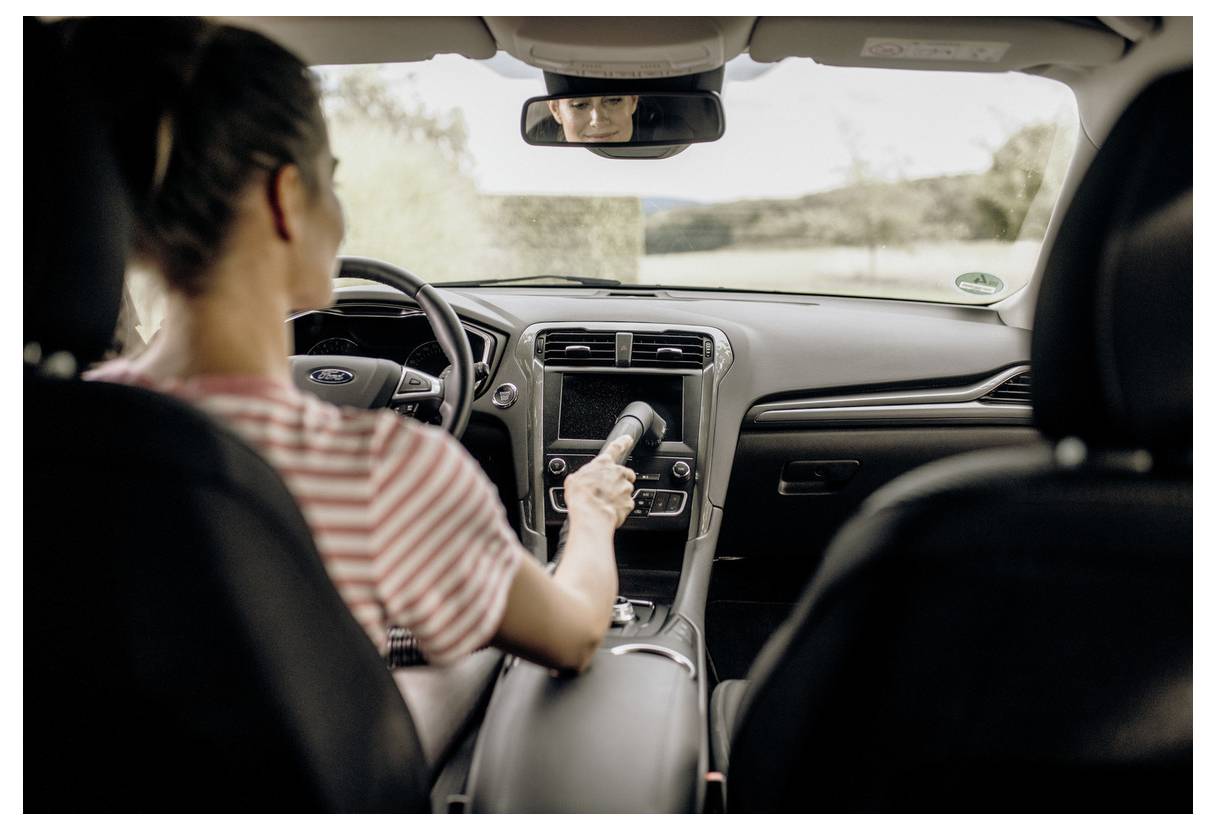 Une personne est assise dans une voiture, en train d'utiliser un petit aspirateur pour nettoyer l'intérieur, alors qu'elle est garée sur une route de campagne.