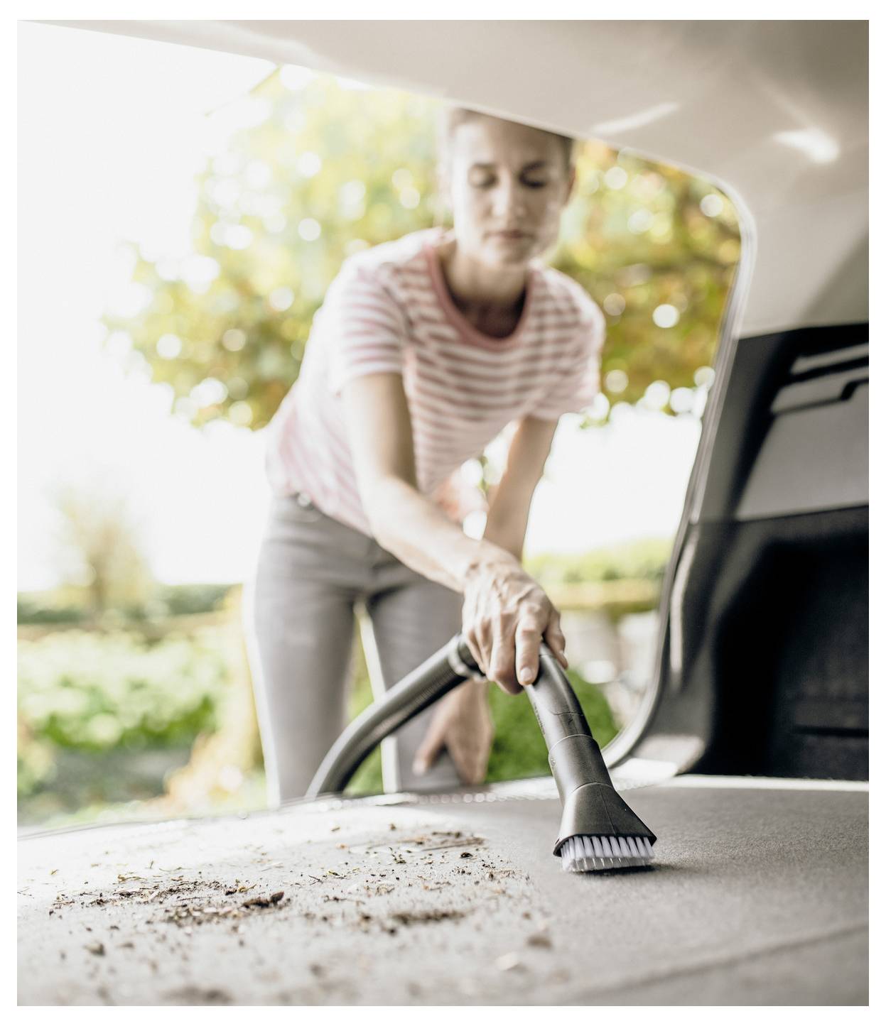 Une personne portant un t-shirt rayé aspire l'intérieur du coffre d'une voiture, en se concentrant sur l'élimination de la saleté et des débris. Des arbres et de la lumière du soleil sont visibles en arrière-plan.
