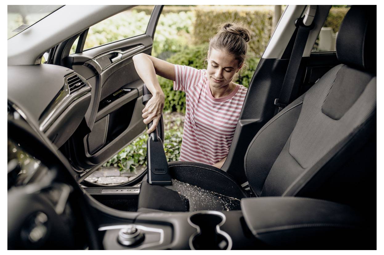 Une femme dans un t-shirt rayé passe l'aspirateur sur le siège passager d'une voiture, en enlevant la saleté. La portière de la voiture est ouverte, et de la verdure est visible à l'extérieur.