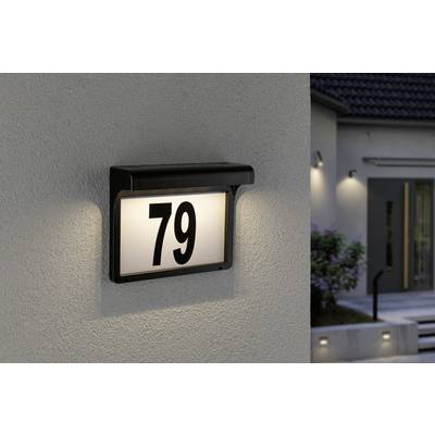 'Plaque de numéro de maison éclairée 79 sur un mur clair ; en arrière-plan, des contours flous d'un bâtiment sont visibles.'