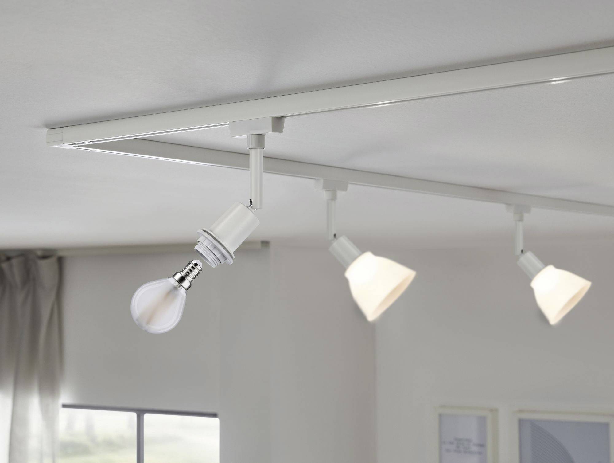 Un plafonnier avec trois spots orientables, dont un sans abat-jour montrant une ampoule à incandescence. Des fenêtres en arrière-plan.