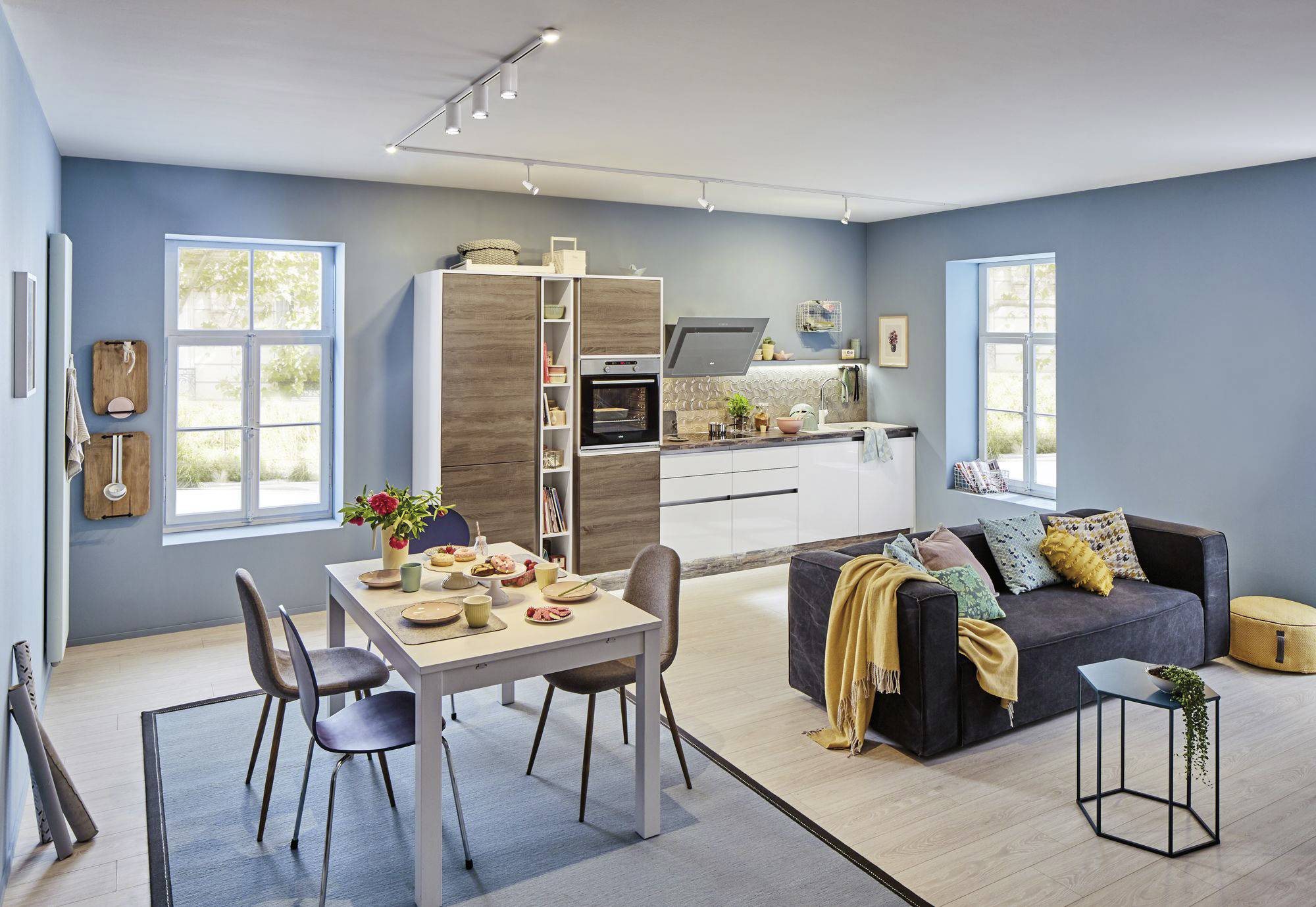 Un salon moderne avec une cuisine ouverte. À gauche, une table à manger est dressée avec de la vaisselle, à droite un canapé sombre avec des coussins et une couverture.